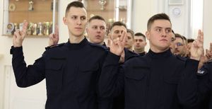 Zdjęcie przedstawia nowych funkcjonariuszy, którzy wypowiadają słowa roty ślubowania. Policjanci mają uniesioną prawą dłoń z wyprostowanymi dwoma palcami.