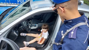 Dziewczynka w radiowozie i stojący obok niej policjant w mundurze.