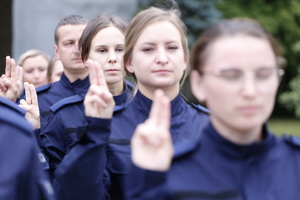 Nowi policjanci z uniesioną prawą dłonią i wyprostowanymi dwoma palcami wypowiadają słowa roty ślubowania.