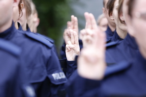 Nowi policjanci z uniesioną prawą dłonią i wyprostowanymi dwoma palcami wypowiadają słowa roty ślubowania.