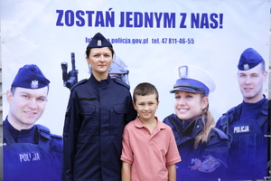 Policjantka z dzieckiem na tle baneru zachęcającego do wstąpienia do policji z napisem zostań jednym z nas!