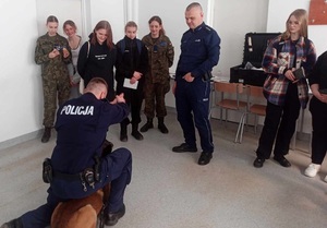 stojący w okręgu uczniowie i na środku policyjny przewodnik z psem w czasie spotkania z młodzieżą