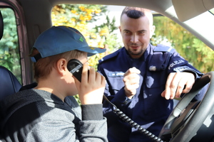 Zdjęcie przedstawia dziecko siedzące w radiowozie które trzyma pilot od sterowania sygnałami uprzywilejowani. koloru szarego. Dziecko zasłoniło nim swoją twarz ubrane jest w czapkę koloru niebieskiego. w tle widoczny jest policjant umundurowany stojący oko radiowozu