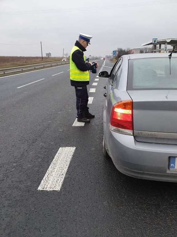 Umundurowany policjant stoi przy pojeździe koloru szarego- dokonuje kontroli drogowej.