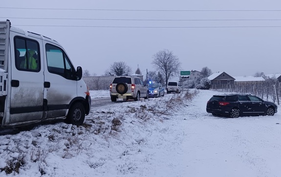 Z lewej strony zdjęcia znajdują się 4 samochody osobowe stojące na jezdni, jeden z samochodów to policyjny oznakowany radiowóz z włączonymi sygnałami świetlnymi koloru niebieskiego. Po prawej stronie na zdjęciu znajduje się pojazd, który wjechał na pobocze.