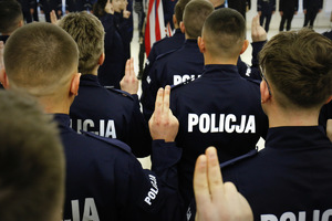 Policjanci ubrani w granatowe mundury z napisami policja ślubują unosząc prawą dłoń z wyprostowanymi dwoma palcami.