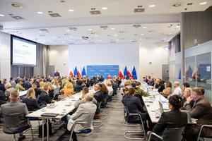 Uczestnicy konferencji zasiadają przy pięciu prostokątnych stołach. Po lewej stronie wyświetlana jest prezentacja. Tło stanowi ścianka MSWiA i flagi Polski i Unii Europejskiej