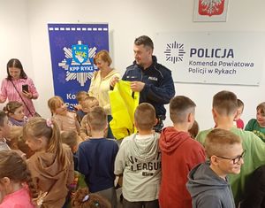 Grupa kilkunastu dzieci podczas zwiedzania budynku Komendy Powiatowej Policji w Rykach w sali odpraw policjant prezentuje kamizelkę odblaskowa policyjną