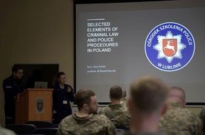 przy mównicy stoi policjant i policjantka, po prawej stronie znajduje się ekran, na którym wyświetlana jest prezentacja multimedialna.