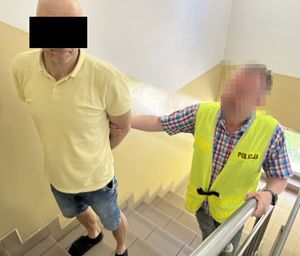 mężczyzna w kajdankach trzymany za ramię przez policjanta po cywilnemu w żółtej odblaskowej kamizelce z napisem policja