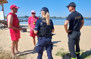 Policjantka ze strażakiem rozmawiają z dwoma ratownikami wodnymi nad zalewem