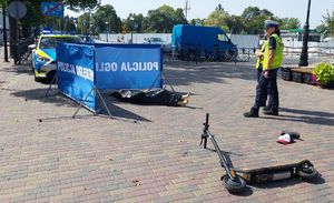 Dwie policjantki stoją w miejscu gdzie przewrócona jest hulajnoga elektryczna znajdują się rozrzucone sportowe buty oraz czapka z daszkiem. Rozłożony jest parawan z napisem &amp;quot;Policja oględziny&amp;quot;, przy którym leży manekin owinięty czarną folią. Za parawanem stoi radiowóz.