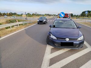 na jezdni zaparkowane samochody. Po prawej stronie na powierzchni wyłączonej z ruchu znajduje się samochód marki Volkswagen, po lewej stronie na pasie jezdni znajduje się samochód marki Renault. Na drugim planie na jezdni znajdują się radiowozy policyjne i wóz strażacki.