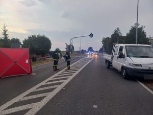 Miejsce wypadku, po lewej stronie znajduje się parawan koloru czerwonego, na środku jezdni stoją strażacy, po parowej stronie samochód marki Fiat koloru białego.