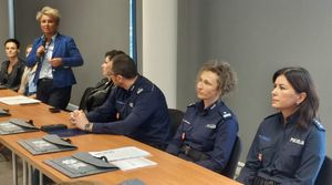 uczestnicy spotkania siedzą przy stole, jedna z kobiet stoi i przemawia przez mikrofon