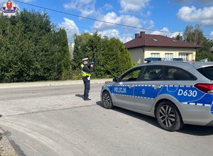 Policjant ruchu drogowego stoi na poboczu mierząc prędkość jazdy kierujących pojazdami. Obok policjanta zaparkowany jest radiowóz.