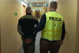 Policjant w odblaskowej kamizelce z napisem Policja prowadzi korytarzem w budynku komendy zatrzymanego mężczyznę
