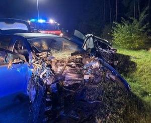 dwa rozbite pojazdy na poboczu drogi, w tle samochód straży pożarnej