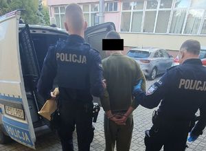dwóch umundurowanych policjantów prowadzi zatrzymanego mężczyznę do radiowozu