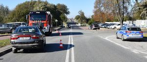 szosa dwujezdniowa, po prawej stronie zaparkowany jest oznakowany radiowóz policyjny, po lewej stronie zaparkowany uszkodzony samochód osobowy i wóz strażacki, w tle samochody zaparkowane na parkingu.