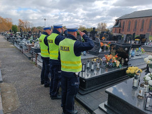 Trzech policjantów umundurowanych z kamizelkami odblaskowymi koloru żółtego odają honor na grobie zmarłego policjanta