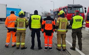 umundurowani funkcjonariusze służb stoją tyłem. W tele znajdują się samochody ciężarowe, radiowóz policyjny i wozy strażackie