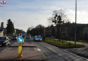 miejsce zdarzenia drogowego - potrącenia pieszej na oznakowanym przejściu dla pieszych, widoczna jest ulica, przejście dla pieszych, oznakowany radiowóz i przejeżdżające samochody