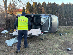 Samochód osobowy leżący na boku z uszkodzeniami po zdarzeniu drogowym oraz odciętym dachem obok stoi umundurowany policjant