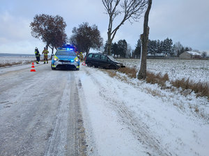 Pojazd marki Opel uszkodzony w wyniku zadurzenia z drzewem obok stoi radiowóz policji na droga jest zaśnieżona i oblodzona