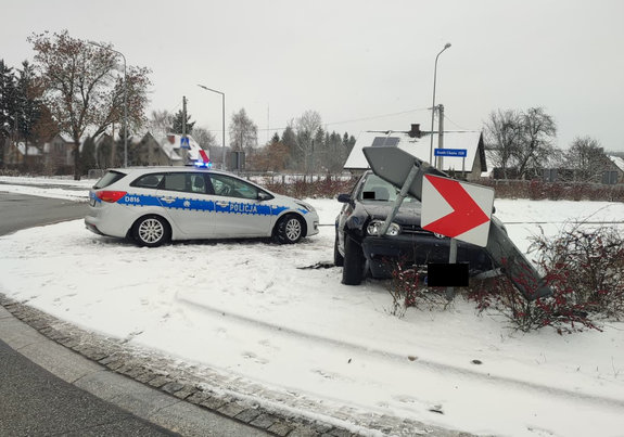 Uszkodzony Volkswagen zawieszony na znakach drogowych na rondzie obok stoi oznakowany radiowóz. teren przykryty śniegiem