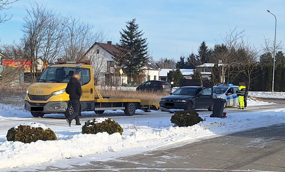 Widoczna jest droga na której stoi laweta koloru żółtego za nią samochód osobowy marki BMW a z zanim radiowóz oznakowany policji z włączonymi sygnałami obok radiowozu stoją dwie osoby w tle zabudowania miejskie oraz inne pojazdy i piesi