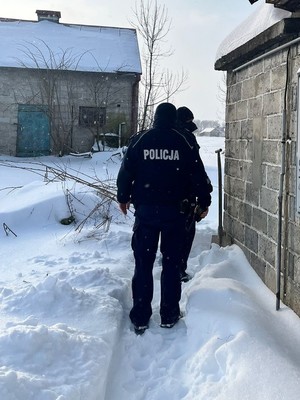 Umundurowani policjanci idą po śniegu przy budynku mieszkalnym