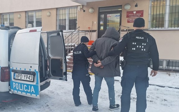 umundurowani policjanci prowadzą zatrzymanego mężczyznę, obok stoi radiowóz, w tle budynek placówki Straży Granicznej