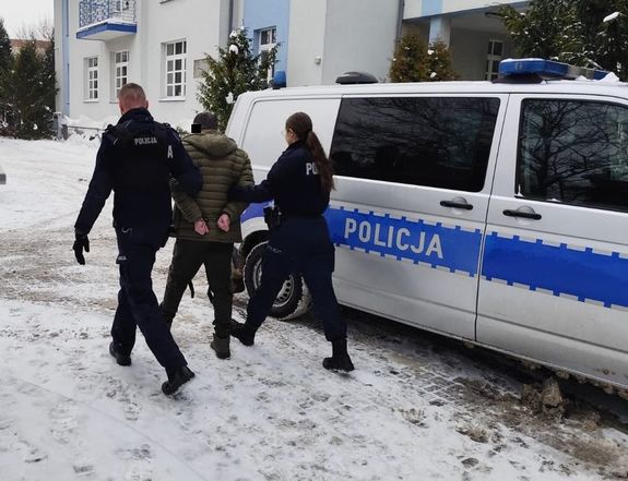Tymczasowo aresztowany 45-latek doprowadzany przez dwóch policjantów do zaparkowanego radiowozu