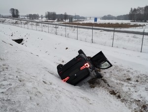 Pojazd marki BMW koloru Czernego  leżący w rowie. Obok stoi radiowóz policji i wystawiony jest znak uwaga wypadek