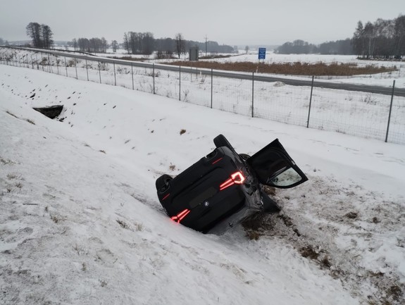 Pojazd marki BMW koloru Czernego leżący w rowie. Obok stoi radiowóz policji i wystawiony jest znak uwaga wypadek