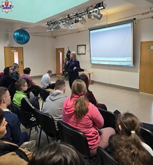 policjantka z mikrofonem stoi przed siedzącymi na krzesłach uczniami podczas spotkania profilaktycznego