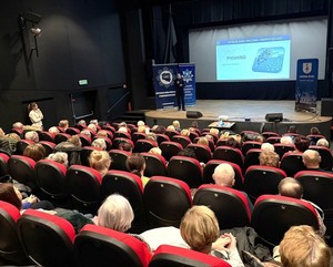 Spotkanie profilaktyczne w sali kina policjanta z seniorami. Funkcjonariusz stoi na scenie seniorzy siedzą w fotelach