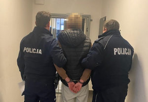 Zatrzymany mężczyzna z założonymi kajdankami jest prowadzony przez dwóch umundurowanych policjantów korytarzem