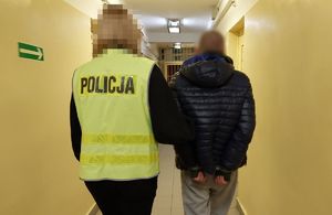 Policjantka w odblaskowej kamizelce z napisem Policja prowadzi korytarzem w budynku komendy zatrzymanego mężczyznę