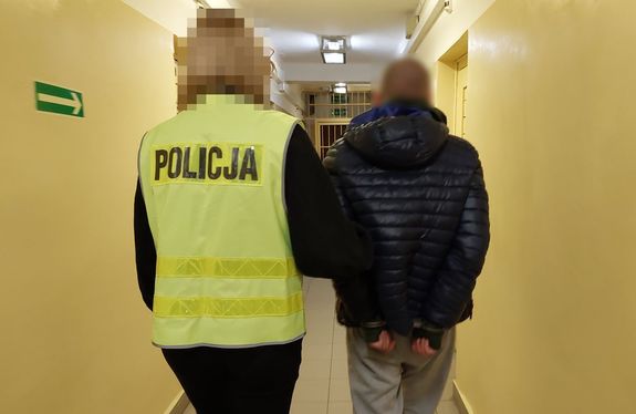 Policjantka w odblaskowej kamizelce z napisem Policja prowadzi korytarzem w budynku komendy zatrzymanego mężczyznę