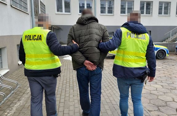 Policjanci w odblaskowych kamizelkach z napisem Policja prowadzą przez parking do radiowozu zatrzymanego mężczyznę