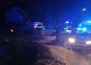 Samochód osobowy leżący na dachu uszkodzony w wyniku zdarzenia drogowego obok na drodze stoją pojazdy służb policji, straży i pogotowia ratunkowego. Policjant wraz z mężczyzną stoi obok radiowozu a strażacy przy srebrnym samochodzie