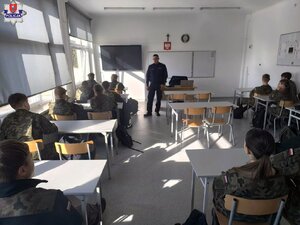 policjant stoi przed siedzącą na krzesłach w ławkach młodzieżą w czasie lekcji