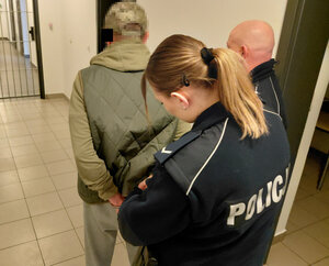 Policjantka zakłada zatrzymanemu mężczyźnie kajdanki na ręce trzymane z tyłu obok stoi jeszcze jedne policjant