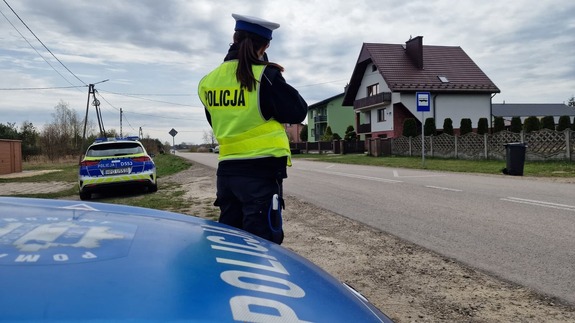 Policjantka mierząca prędkość ręcznym miernikiem prędkości obok jezdni, na pierwszym planie widoczna maska radiowozu z napisem POLICJA