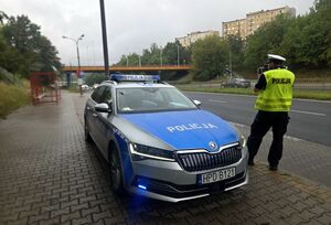 obok radiowozu stoi policjant i mierzy prędkość pojazdów