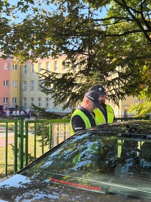 dwóch umundurowanych policjantów stoi przy samochodzie osobowym, w tle jest szkoła i dzieci