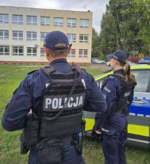 dwoje umundurowanych policjantów stoi przy oznakowanym radiowozie